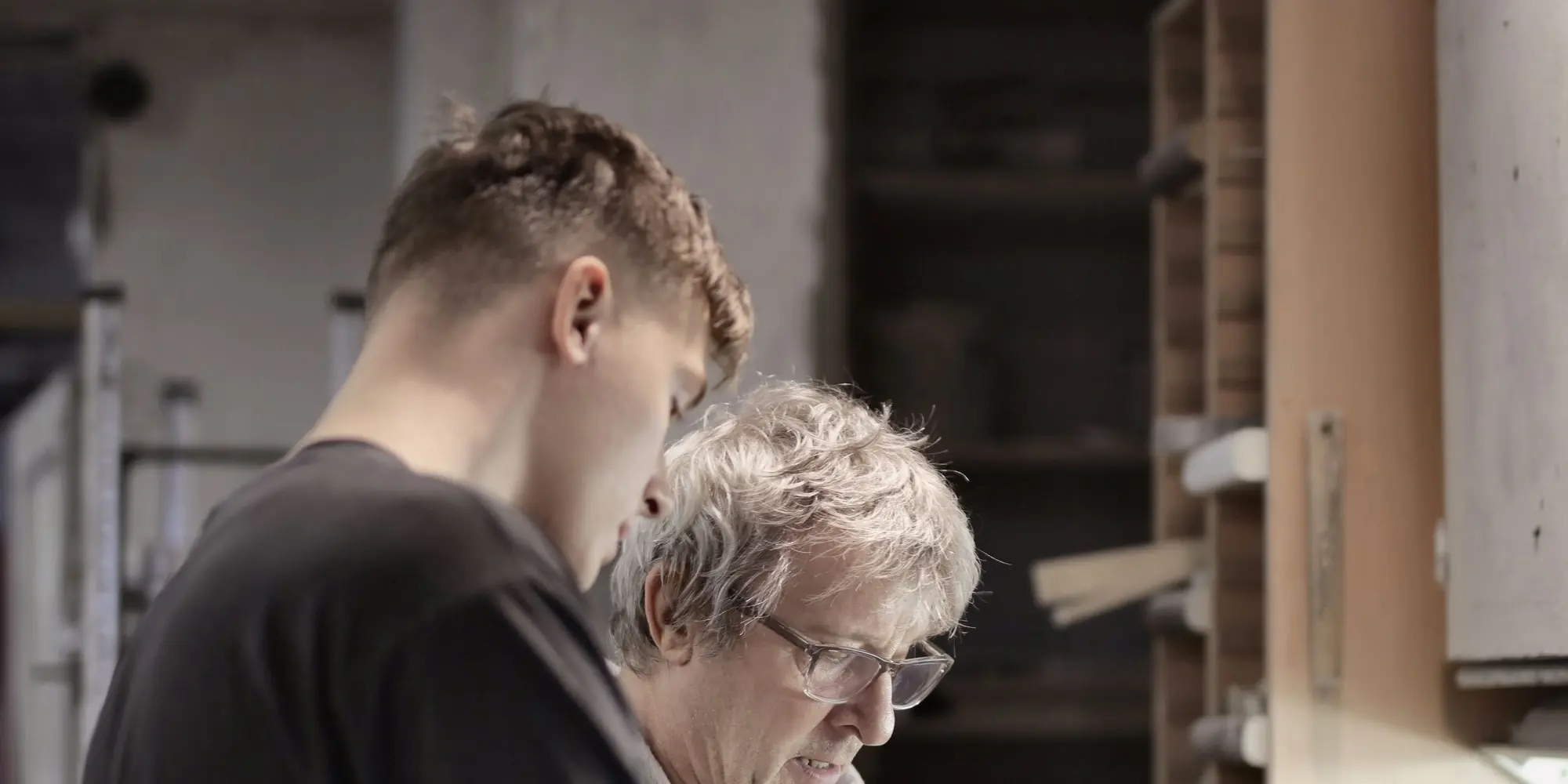 A young apprentice and an experienced tradesman working side by side at a workshop bench.